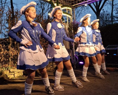 Vier Tänzerinnen in blauen Kostümen führen eine Aufführung im Freien auf.